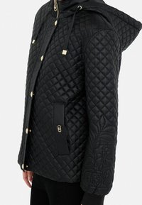 Chaqueta negra acolchada con capucha, herrajes dorados y detalle estampado en la manga. Incluye un bolsillo lateral con un acento de logo.