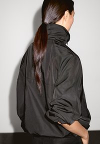Donna con i lunghi capelli scuri raccolti, che indossa una giacca nera larga con le maniche arrotolate, mostrata in vista laterale-posteriore su uno sfondo semplice.