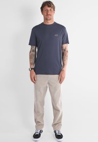 T-shirt en coton bleu marine avec un petit logo crème, associé à un pantalon en velours côtelé beige clair et des baskets noires avec des lacets blancs.