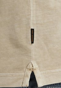 Chemise en coton beige avec un tissu texturé. Présente une étiquette de logo "PME LEGEND" près de l'ourlet. Comprend un détail d'ourlet fendu pour le style.