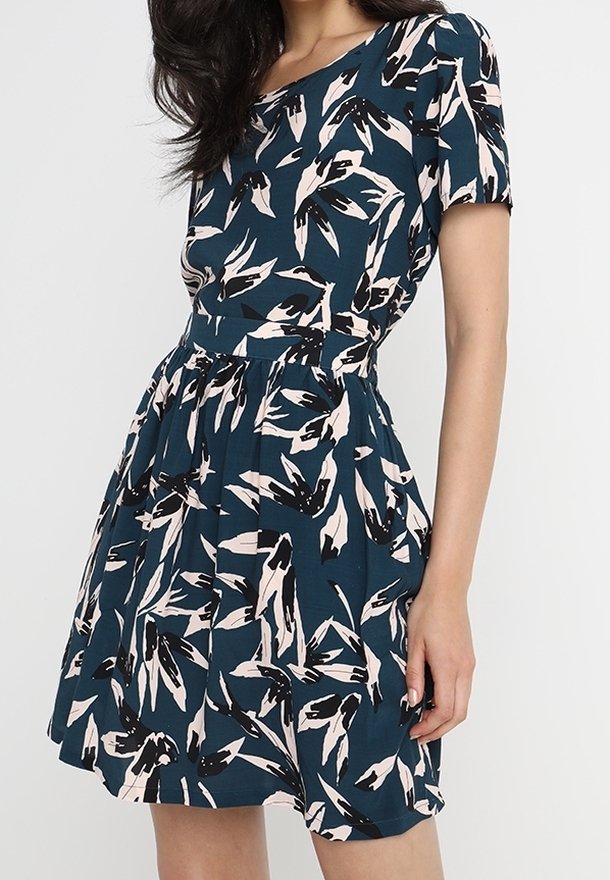 Femme portant une robe à manches courtes, longueur genou, avec un motif abstrait de feuilles bleu foncé et blanc.