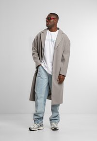 Gray long coat over a white t-shirt with text, light blue jeans, and gray sneakers with reflective accents. Red sunglasses complete the outfit.