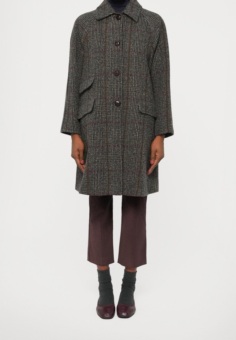 Personne portant un manteau marron à carreaux longueur genou avec de gros boutons, pantalon court foncé, chaussettes grises et chaussures bordeaux, debout sur fond blanc.