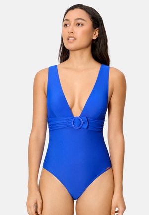 Mujer con un traje de baño de una pieza azul con escote en V profundo y un detalle circular de hebilla en la cintura, posando contra un fondo liso.