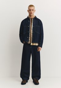 Temno modra denim jakna z dvema žepoma na prsih, nošena preko svetle karo srajce. Ohlapne temno modre kavbojke in rjavi semiš čevlji.