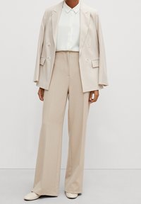 Blazer beige cintré à double boutonnage porté sur une chemise blanche, associé à un pantalon beige taille haute à jambes larges et des mocassins de couleur claire.