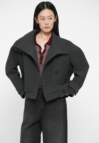 �Ženska z dolgimi črnimi lasmi nosi temno sivo kratko jakno preko burgundske srajce z gumbi in črnimi hlačami visoke pasu, stoji z rokama v žepih.