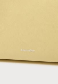 Yellow textured leather wallet with embossed white "Calvin Klein" logo on the front. Minimalist design, clean lines, smooth finish.