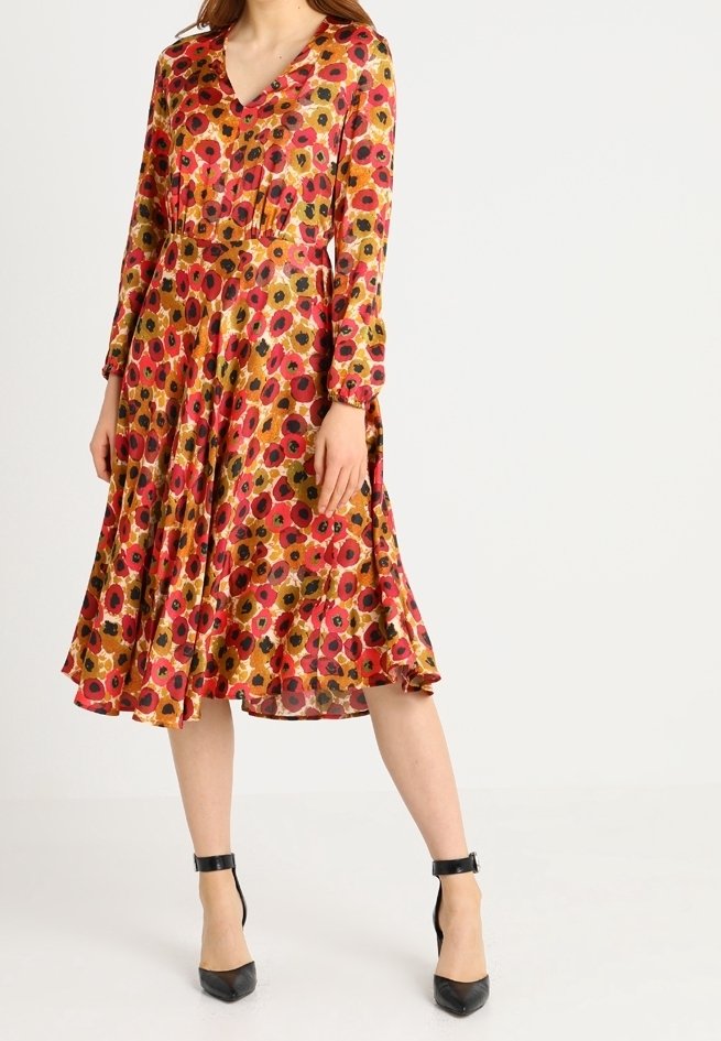 Femme portant une robe manches longues, longueur genou, avec un motif floral rouge, orange et noir, associée à des talons noirs à bride cheville.