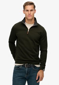 Joven vestido con un suéter de cremallera hasta el cuarto en color verde oscuro sobre una camisa beige, jeans azules, cinturón negro y reloj de pulsera, de pie frente a un fondo blanco.