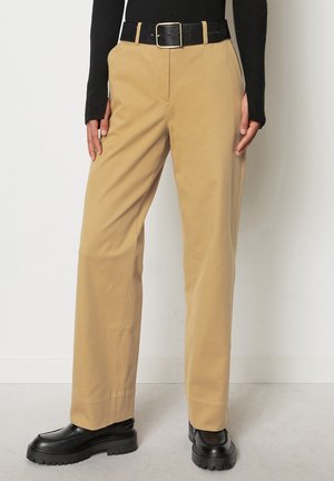 Beige wijde broek gemaakt van gladde stof, met een zwarte riem met een gouden gesp, gecombineerd met zwarte schoenen met een dikke zool.