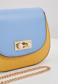 Bolso bicolor de cuero azul y amarillo con un cierre dorado que presenta dos perlas blancas. Incluye una correa de cadena dorada.