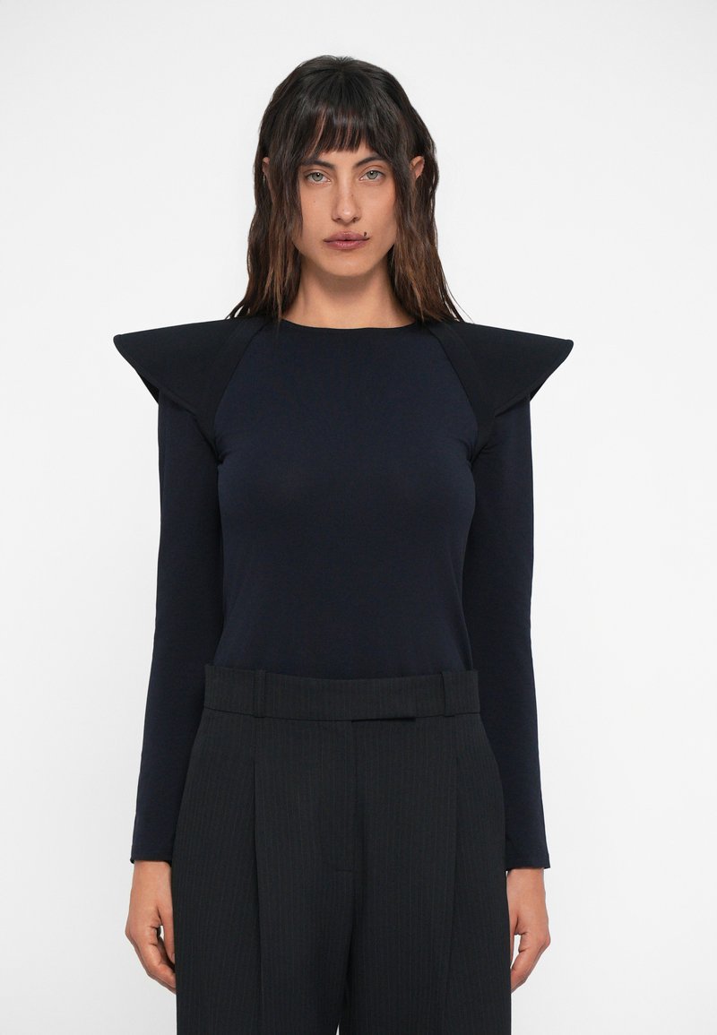 Camisola de manga longa azul-marinho com ombros distintivos, feita de um tecido suave. Apresenta uma silhueta ajustada combinada com calças de riscas de cintura alta.