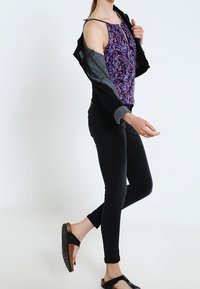Fioletowa kwiecista bluzka bez rękawów z dekoltem w kształcie łezki, czarne dżinsy oraz czarne sandały na drewnianej, platformowej podeszwie. Czarna kurtka zarzucona na ramię.