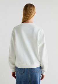 Felpa bianca con scollatura a girocollo a coste e maniche lunghe, caratterizzata da una vestibilità comoda e una texture morbida. Indossata con una gonna di jeans, parzialmente visibile.