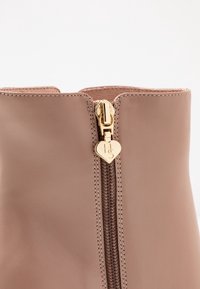 Brązowy skórzany ботинок sięgający kostki z złotym zamkiem błyskawicznym i uchwytem w kształcie serca. Zamek biegnie pionowo wzdłuż przodu, z widocznymi detalami szwów.