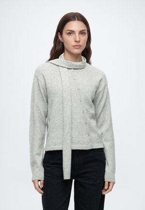 Femme aux cheveux bruns raides porte un pull gris clair boutonné avec un détail écharpe et un pantalon noir, debout devant un fond blanc uni.