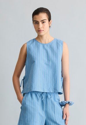 Junge Frau trägt ein hellblaues ärmelloses gestreiftes Oberteil und passende Shorts mit Kordelzug sowie ein passendes Haargummi am Handgelenk.