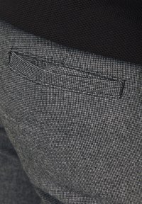 Grauer, texturierter Stoff von Hosen mit einem subtilen Karomuster, wobei eine Paspeltasche auf der Rückseite sichtbar ist.