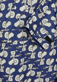 Blauwe stof met herhaalde zwart-witte illustraties van Snoopy. Soepele textuur met een glanzende afwerking; voorzien van drukknoopjes en klittenbandaccenten.