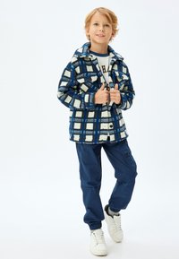 Chaqueta de cuadros forrada de forro polar en azul y blanco, con dos bolsillos frontales, llevada sobre una camiseta gráfica, combinada con joggers marinos y zapatillas blancas.