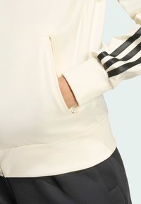 Chaqueta blanca con cremallera que presenta acentos en negro con tres rayas en las mangas, puños acanalados y bolsillos laterales con una textura de tela suave.