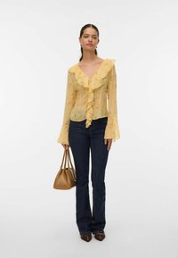 Blouse jaune transparente avec un col froncé et des manches évasées, associée à un jean évasé bleu foncé et un sac à main en cuir marron.