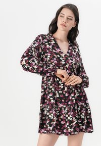 Robe noire à fleurs avec manches longues ; présente un col en V et des motifs de fleurs multicolores en rose, blanc et violet. Texture de tissu douce.