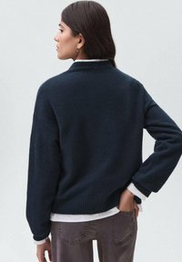 Kvinna med mörkt, rakt hår, iförd en marinblå tröja över en vit skjorta och grå jeans, stående med ena handen i bakfickan.