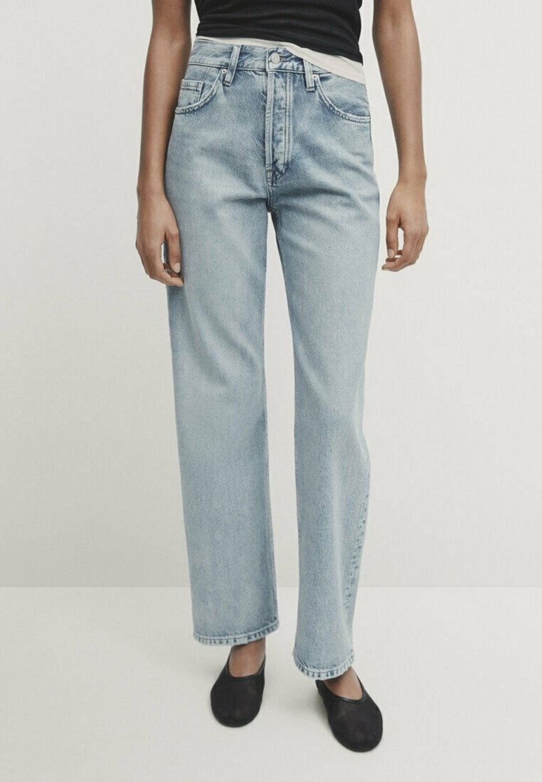 Osoba ubrana w jasnoniebieskie luźne dżinsy, czarną bluzkę i czarne buty typu slip-on, stojąca na tle białego, jednolitego tła.