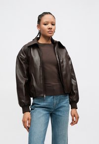 Καφέ δερμάτινο μπουφάν τύπου μπούντερ με φερμουάρ εμπρός, μεγάλο γιακά και ριμπ μανσέτες, συνδυασμένο με καφέ τοπ και ανοιχτό μπλε τζιν με φθορές.