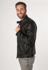 Schwarze Lederjacke mit hohem Kragen, Reißverschluss vorne und strukturierten Schulterakzenten, kombiniert mit hellgrauen Jeans.