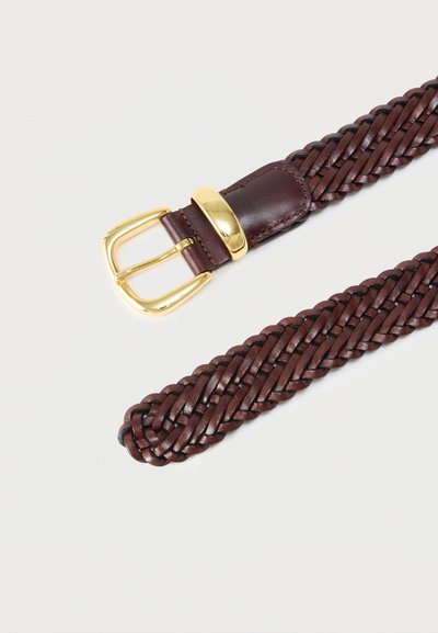 Πλεγμένη ζώνη από σκούρο καφέ δέρμα με χρυσή μεταλλική αγκράφα και λεπτομέρεια θηλιάς σε ανοιχτό φόντο.