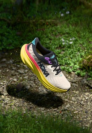 Lys beige løbesko med sorte snørebånd, gul sål og farverige accenter, der svæver over grus og græs udendørs.
