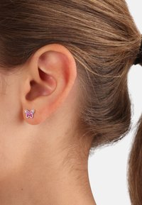 Orecchino a forma di farfalla rosa con accenti brillanti, realizzato in metallo, posizionato su un primo piano di un orecchio con capelli castani tirati indietro.