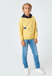 Sudadera amarilla con capucha azul marino, bolsillo frontal y etiqueta azul; combinada con jeans de mezclilla azul claro y zapatillas blancas. Textura suave.