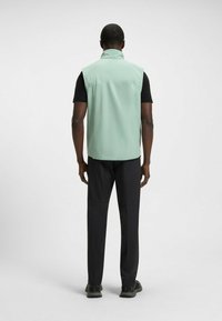 Ärmelloses mintgrünes Weste mit hohem Kragen, glatter Textur und Rückenschlitz, kombiniert mit schwarzen Hosen und dunklem Schuhwerk.