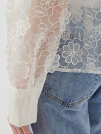 Blusa bianca trasparente con ricami floreali sulle maniche, con polsini bianchi solidi con bottoni e jeans in denim visibili sullo sfondo.