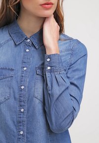 Chemise en denim bleu clair avec un col pointu, un devant à boutons, deux poches poitrine et des poignets froncés. Le tissu présente une texture subtile.