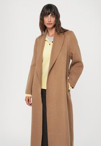Manteau long marron en tissu de laine douce, avec un col cranté et un design à l'avant ouvert. Porté par-dessus un haut jaune pâle.