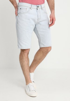Mann trägt hellblaue Jeansshorts, weiße Sneaker und ein pinkfarbenes T-Shirt, steht mit einer Hand in der Tasche.
