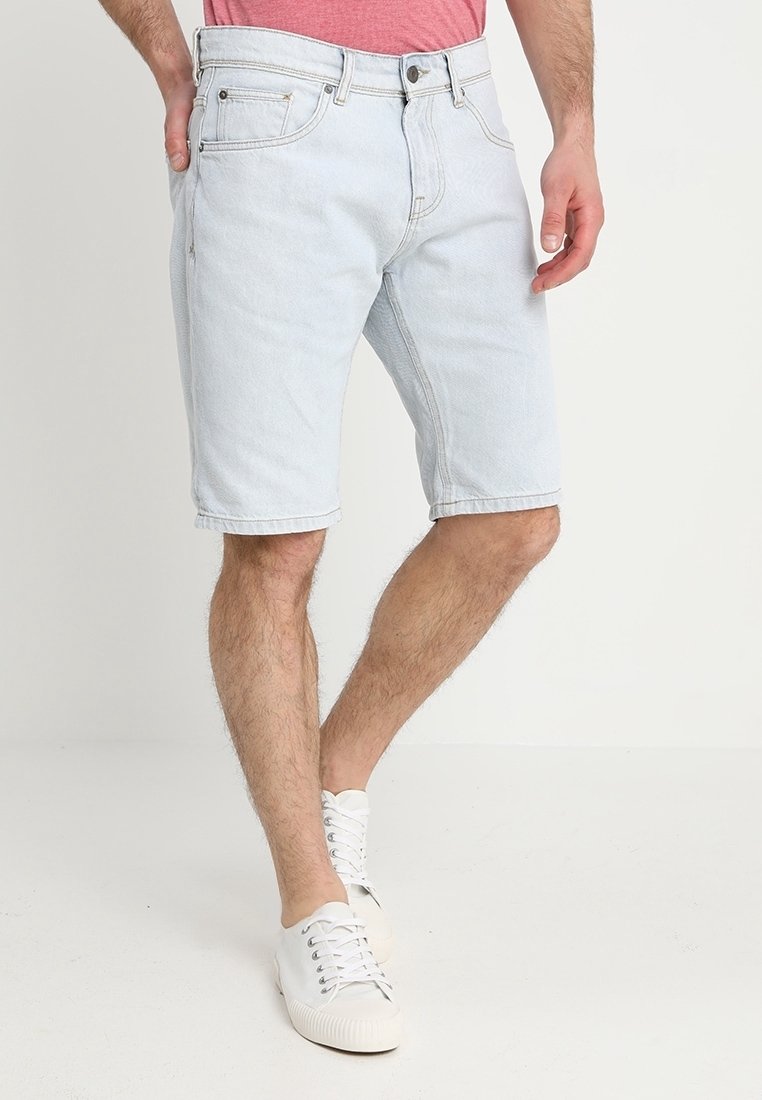 Homme portant un short en jean bleu clair, des baskets blanches et un t-shirt rose, debout avec une main dans la poche.
