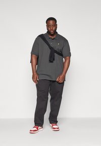 Polo shirt grigio scuro con logo giallo, pantaloni neri e sneakers rosso e bianche. Un maglione nero è appoggiato sulla spalla.