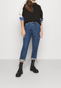 Sudadera negra con detalles en blanco y azul, mangas azules remangadas, combinada con vaqueros azules de talle alto y botas negras de plataforma con cordones.