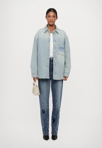 Gaiši zils pārspīlēts džinsu jakas ar krūšu kabatu, apvienots ar tumši zilām džinsām, kas rotātas ar pērļu akcentiem un miers simbolu rakstiem.