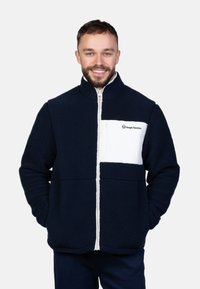 Marineblaue Fleecejacke mit einer weißen Brusttasche und Reißverschluss. Verfügt über einen hohen Kragen und strukturierten Stoff, der für Wärme und Komfort sorgt.