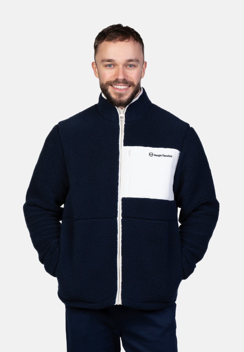 Marineblaue Fleecejacke mit einer weißen Brusttasche und Reißverschluss. Verfügt über einen hohen Kragen und strukturierten Stoff, der für Wärme und Komfort sorgt.