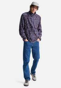 Fleece jacket in dark purple with textured white abstract patterns, high collar, front zipper, and two side pockets. Worn with blue jeans.