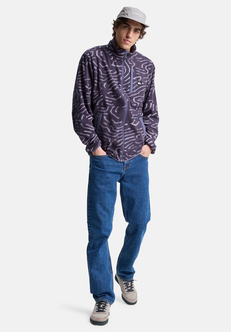 Fleece jacket in dark purple with textured white abstract patterns, high collar, front zipper, and two side pockets. Worn with blue jeans.