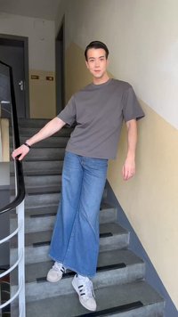 Νέος άνθρωπος με γκρι μπλουζάκι και φαρδιά μπλε τζιν που στηρίζεται σε κιγκλίδωμα σκάλας σε μπεζ και γκρι σκάλες.